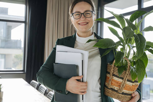 Smiling businesswoman holding potted plant and file folder in office - OSF02352