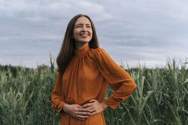 Cheerful woman with hands on hip standing by corn crops at field - TOF00203