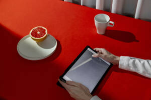 Hände einer Frau, die einen Tablet-PC in der Nähe einer Grapefruit und einer Teetasse an einem roten Tisch benutzt - MBLF00220
