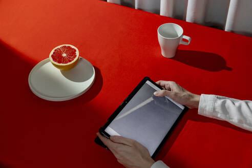 Hands of woman using tablet PC near grapefruit and tea cup at red table - MBLF00220