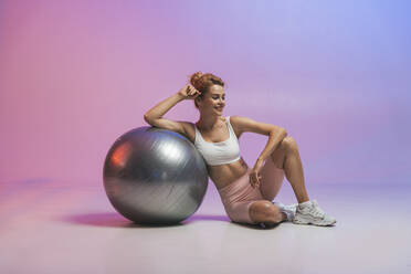 Lächelnde Frau, die sich auf einen Fitnessball stützt, der vor einem zweifarbigen Hintergrund sitzt - OIPF03670