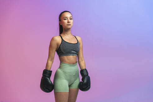 Confident young woman standing with black boxing gloves against colored background - OIPF03695