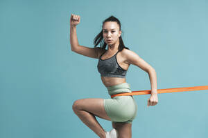 Confident young woman exercising with resistance band against blue background - OIPF03758