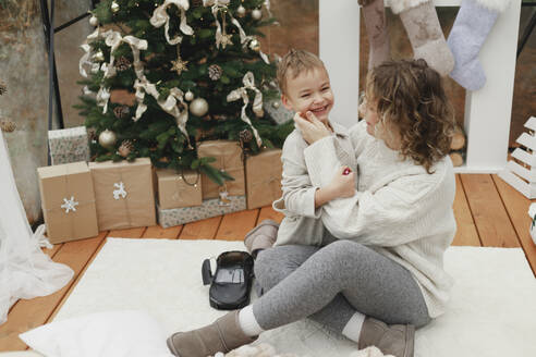 Glückliche Mutter umarmt Sohn auf Teppich in der Nähe von Weihnachtsbaum zu Hause - EHAF00163