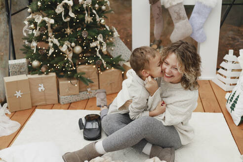 Junge küsst lächelnd Mutter sitzt auf Teppich in der Nähe von Weihnachtsbaum zu Hause - EHAF00164