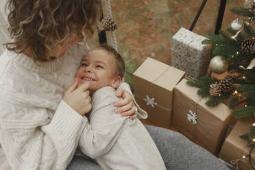 Liebevolle Mutter umarmt Sohn in der Nähe von Weihnachtsgeschenken zu Hause - EHAF00170