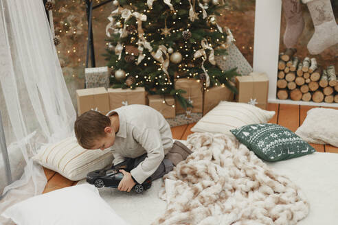 Junge spielt mit Spielzeugauto auf Teppich in der Nähe von Weihnachtsbaum zu Hause - EHAF00185