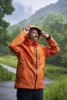 Glücklicher Mann mit orangefarbener Jacke und vor einem Berg stehend - VEGF06230