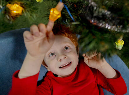 Niedlicher Junge spielt mit Weihnachtsbeleuchtung unter dem Baum - MBLF00231