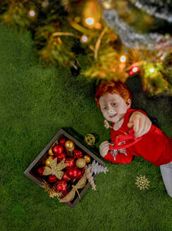 Glücklicher rothaariger Junge spielt mit Ornament am Weihnachtsbaum auf grünem Teppich - MBLF00234