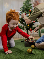 Redhead boy playing with bauble near grandfather decorating Christmas tree at home - MBLF00235