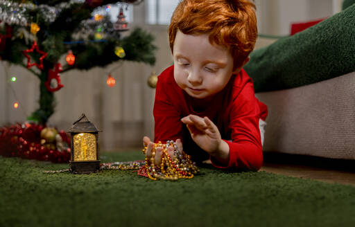 Süßer rothaariger Junge spielt mit Weihnachtsschmuck auf grünem Teppich zu Hause - MBLF00236