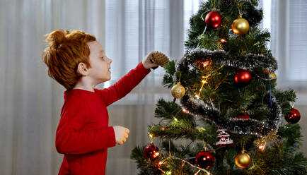 Redhead boy decorating Christmas tree with pine cone at home - MBLF00238