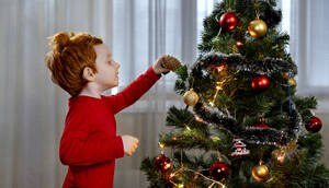 Rothaariger Junge schmückt Weihnachtsbaum mit Tannenzapfen zu Hause - MBLF00238