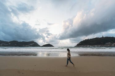 Junge Frau, die am Strand von Playa de la Concha bei bewölktem Himmel spazieren geht - DAMF01250