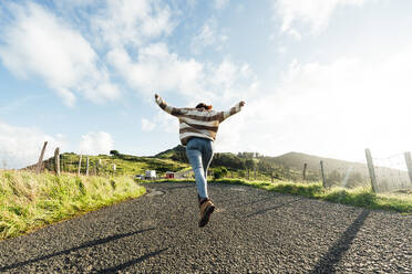 Young woman with arms outstretched running on road under cloudy sky - DAMF01268