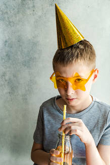 Ein Junge mit einem glitzernden Partyhut und einer gelben, sternförmigen Brille trinkt mit einem gestreiften Strohhalm aus einer Glasflasche vor einem neutralen Hintergrund. - ADSF51809