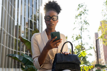 Beautiful African American businesswoman messaging over smartphone while sitting with black purse in urban city against building - ADSF51938