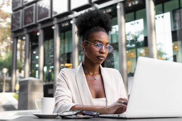 Fokussierte junge afroamerikanische Geschäftsfrau mit lockigem Haar, die eine Brille trägt und an einem Laptop arbeitet, während sie in einem Straßencafé sitzt - ADSF51941