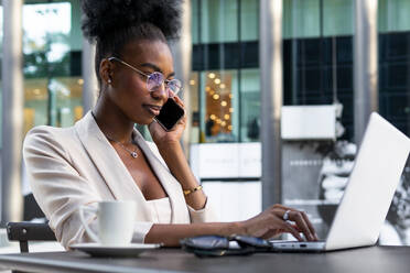 Side view of smiling young African American businesswoman having business call over mobile phone and using laptop at sidewalk cafe - ADSF51942