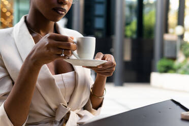 Anonymous crop African American female professional with eyeglasses holding coffee cup while sitting at sidewalk cafe - ADSF51943