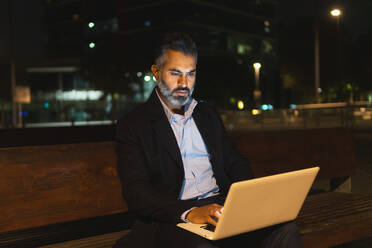 Seriöser bärtiger Geschäftsmann im Anzug surft mit Netbook im Internet, während er abends auf einer Holzbank vor beleuchteten Gebäuden einer modernen Stadt sitzt - ADSF51951