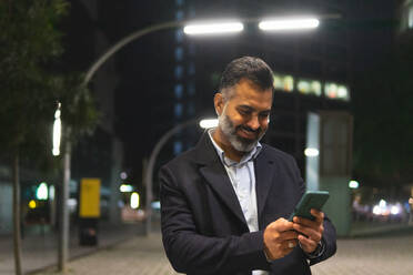 Lächelnde bärtigen indischen Mann im Anzug stehen auf der Straße mit Straßenlaternen und SMS mit Handy gegen beleuchtete Gebäude der Stadt - ADSF51952