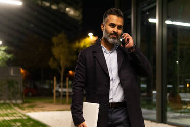Focused Indian entrepreneur in classy outfit holding laptop and talking on smartphone after successful business meeting - ADSF51961