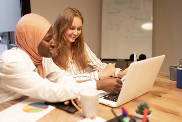 Lächelnde multiethnische junge Kolleginnen, die am Schreibtisch sitzen, einen Laptop benutzen und an einem Projekt in einem modernen Büro arbeiten, während sie eine Geschäftsstrategie diskutieren - ADSF51971