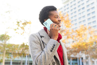 Smiling African American female speaking on mobile phone while standing against blurred modern buildings in city street - ADSF51983