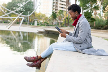 Seitenansicht einer lächelnden Afroamerikanerin in Oberbekleidung, die in der Nähe eines Teiches sitzt und ein elektronisches Buch liest, während sie sich in einem Park am Wochenende ausruht - ADSF51991