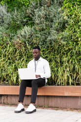 Ganzkörperdarstellung eines jungen afroamerikanischen Geschäftsmannes in formeller Kleidung, der auf einer Bank vor einer Hecke sitzt und über ein Netbook aus der Ferne arbeitet - ADSF51994