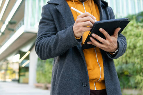 Ein Geschäftsmann in eleganter Freizeitkleidung benutzt aufmerksam ein digitales Tablet, das vor einem modernen Gebäude steht. - ADSF52000