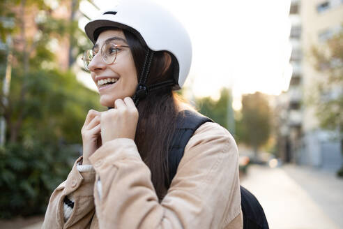 Eine fröhliche junge Frau ist zu sehen, wie sie ihren Schutzhelm befestigt, bereit für eine Fahrt durch die Stadt. - ADSF52003