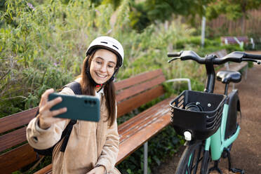 Eine lächelnde junge Frau macht ein Selfie mit ihrem Smartphone, während sie einen Schutzhelm trägt und neben ihrem Fahrrad in einer üppigen Parklandschaft steht. - ADSF52006
