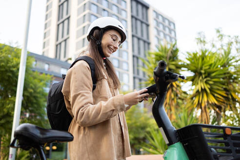 Eine lächelnde junge Frau in Freizeitkleidung und mit Helm benutzt ihr Smartphone, während sie neben einem Elektroroller in einer städtischen Umgebung steht. - ADSF52008