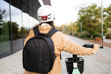Ein junger Stadtpendler mit Helm und Rucksack steht an einem Gebäude, in der Hand ein Smartphone und ein Fernglas. - ADSF52009