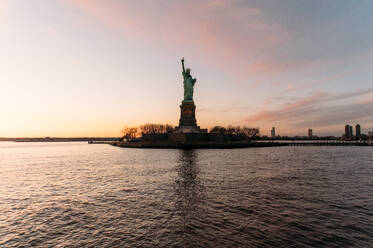 Malerischer Blick auf die Freiheitsstatue am Ufer eines ruhigen Flusses gegen den bewölkten Himmel bei Sonnenuntergang in New York - ADSF52038