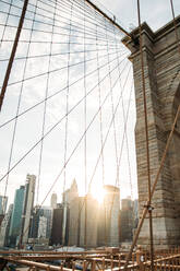 Sunlit view of the Brooklyn Bridge's intricate cable design, with the Manhattan skyline in the background during sunset. - ADSF52040