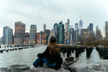 Eine Frau sitzt in der Dämmerung auf einem Felsen mit Blick auf die Skyline von Manhattan. - ADSF52044