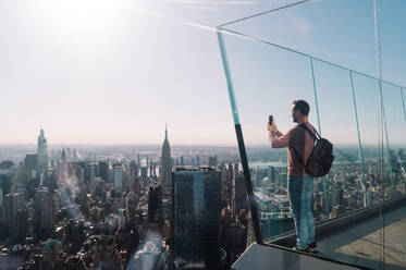 Ein Mann in legerer Kleidung und mit einem Rucksack macht mit seinem Smartphone ein Foto von der weitläufigen Skyline von Manhattan, die in sanftes Sonnenlicht getaucht ist. - ADSF52061