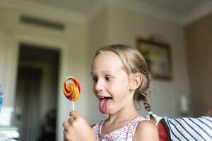 Ein fröhliches junges Mädchen mit geflochtenen Haaren schaut sich in einem hellen Haus einen großen, bunten Lutscher an. - ADSF52070
