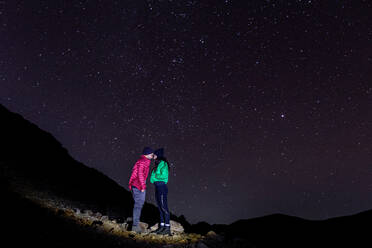 Seitenansicht von anonymen Reisenden Paar küssen und in warmer Kleidung, während stehend auf Hochland felsigen Klippe im Licht unter spektakulären Landschaft der Sternenhimmel in der Nacht Zeit - ADSF52071