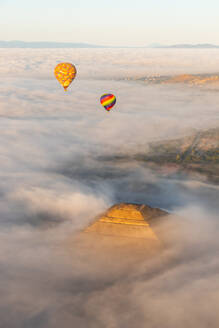 Heißluftballons, die bei Sonnenaufgang über einer dichten Wolkendecke aufsteigen, wobei sich die goldenen Farbtöne der Morgensonne auf der Spitze der Sonnenpyramide in Teotihuacan, Mexiko, spiegeln - ADSF52072