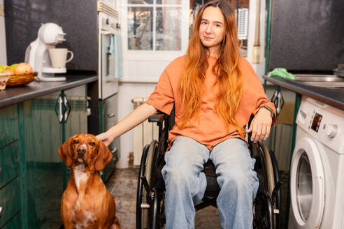 A woman in a wheelchair pets her dog in a cozy home kitchen, displaying companionship and accessibility. - ADSF52082