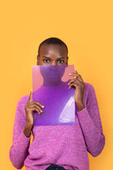 Portrait of African American young woman in pink sweater holding transparent plastic while standing looking at camera against yellow background - ADSF52126