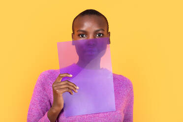 Porträt einer afroamerikanischen jungen Frau in einem rosa Pullover, die eine durchsichtige Plastikfolie hält und in die Kamera schaut, vor einem gelben Hintergrund - ADSF52128