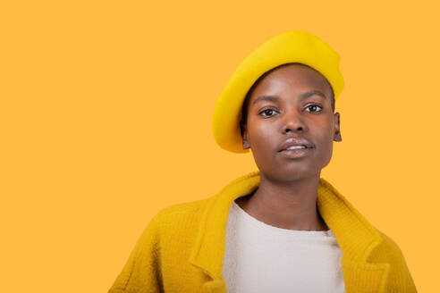 Nachdenkliche, modische afroamerikanische junge Frau, die eine flache Mütze und einen Mantel trägt und in die Kamera schaut, vor einem gelben Hintergrund - ADSF52131