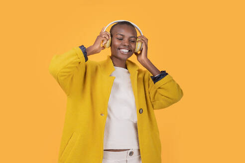 Fröhliche afroamerikanische junge Frau, die in ihrer Jacke nach unten schaut und über drahtlose Kopfhörer vor gelbem Hintergrund Musik hört - ADSF52132
