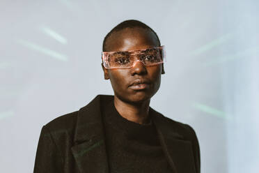 Porträt einer selbstbewussten jungen, mutigen afroamerikanischen Frau mit einer intelligenten, futuristischen VR-Brille und einem langen schwarzen Trenchcoat, die in die Kamera blickt, vor einem mit Neonlicht beleuchteten Hintergrund - ADSF52137